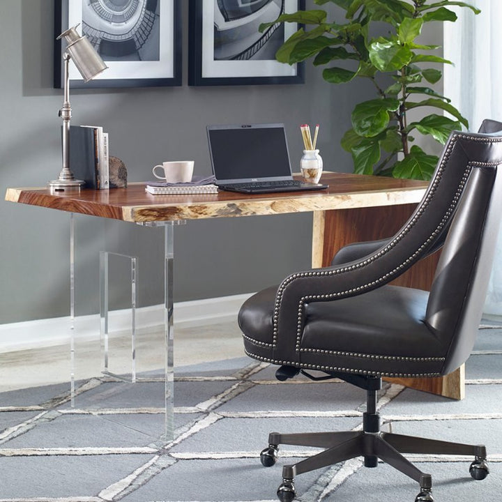 A stylish home office setup featuring the Fairfield Chair Live Edge Natural Guanacaste Waterfall Desk 54" with an acrylic base. On the desk, there's a laptop, a cup of coffee, and a desk lamp. A black leather office chair with nailhead trim faces the desk. In the background, there are framed pictures and a large plant.