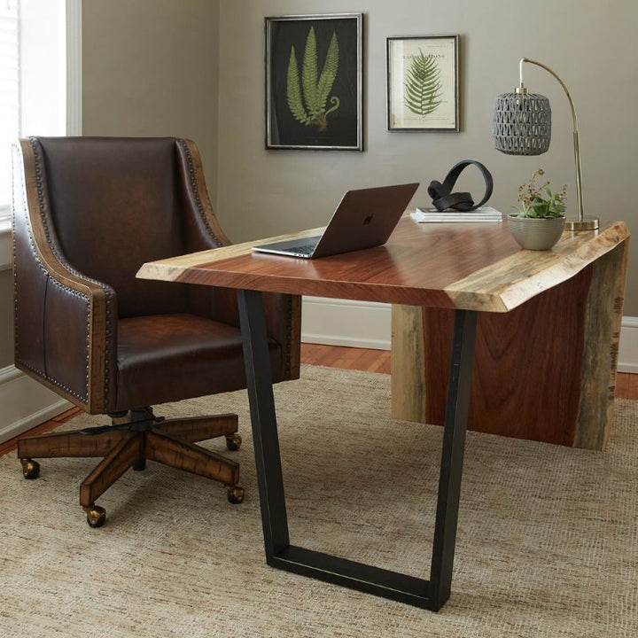 A modern office setup featuring the Live Edge Natural Guanacaste Waterfall Desk 54" with a trapezoid base from Fairfield Chair, paired with a brown leather office chair adorned with nailhead trim. A laptop rests on the desk, while the room is decorated with framed pictures of ferns, a lamp, a sculpture, and a plant.