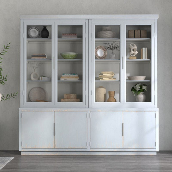 A Sarreid Maldives 4 Door Bookcase in Eros Blue, featuring a gray wooden frame and glass doors, showcases various decor items on its shelves, including vases, bowls, books, and framed pictures. Below the glass cabinets are solid wood doors for additional storage. A green potted plant is partially visible on the left side of this reclaimed oak bookcase.