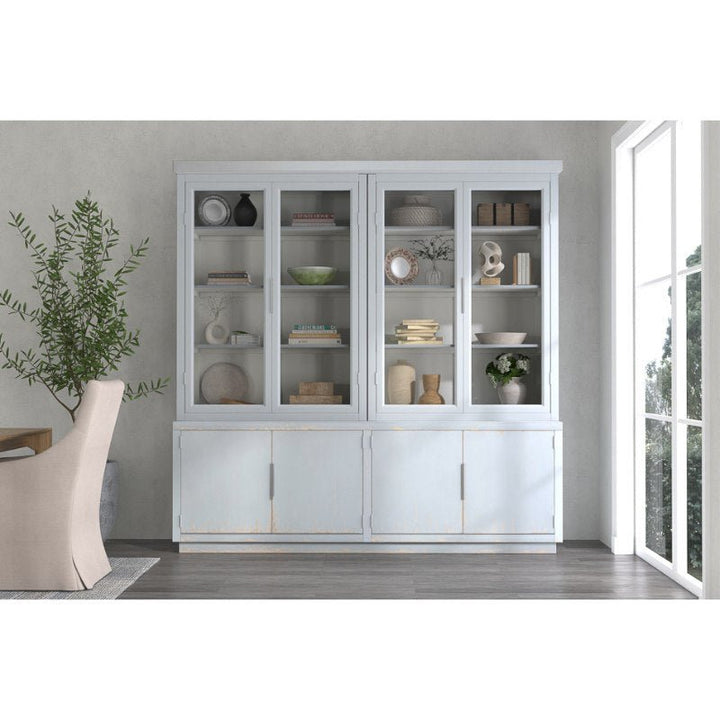 A striking wooden display cabinet, painted in Eros Blue and reminiscent of the Maldives 4 Door Bookcase by Sarreid, stands against a light-colored wall. The glass doors reveal an array of decorative items, including bowls, books, and vases. The lower section features closed cabinets made from reclaimed oak. Through the room, a dining table and chairs are partially visible.