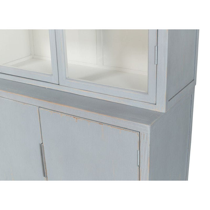 A partial view of a two-tiered, light blue wooden cabinet. The upper section has glass-paneled doors, while the lower section features solid doors. With its distressed Eros Blue finish and slight wear and tear for a vintage look, it exudes the elegant charm of the Maldives 4 Door Bookcase by Sarreid.