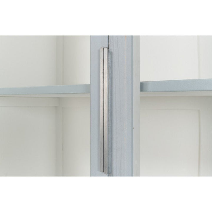 Close-up view of a gray cabinet handle on a light-colored cabinet door. The handle is long, slender, and metallic with a polished finish, contrasting beautifully with the Eros Blue backdrop. Centrally located on the vertical edge of the door, it reveals shelves in what appears to be part of Sarreid's Maldives 4 Door Bookcase in Eros Blue.