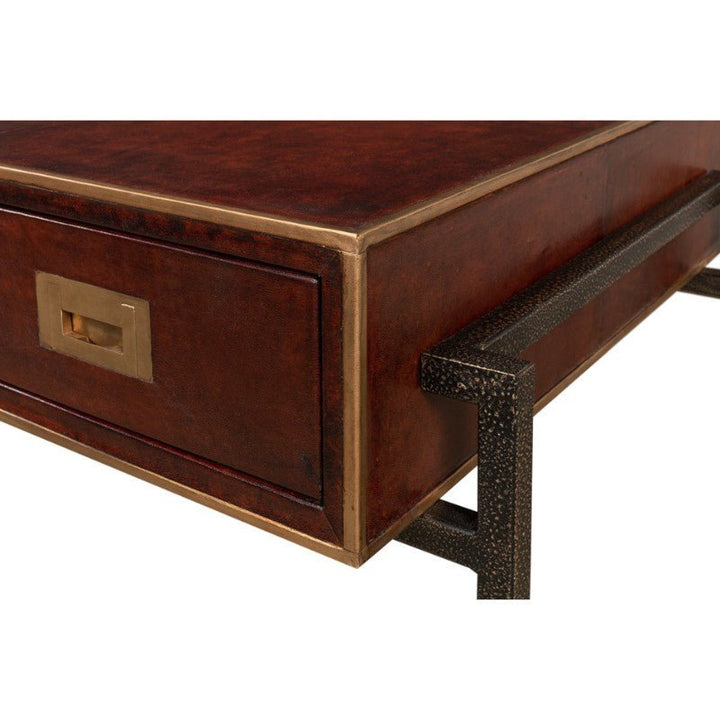 A close-up of a corner of the Sarreid Old Leather Desk, featuring a dark brown leather surface with brass accents. This luxurious piece includes a rectangular brass handle on one of its drawers and stands on textured black iron legs. The polished, hand-colored leather top adds a touch of transitional style to this 49" desk.