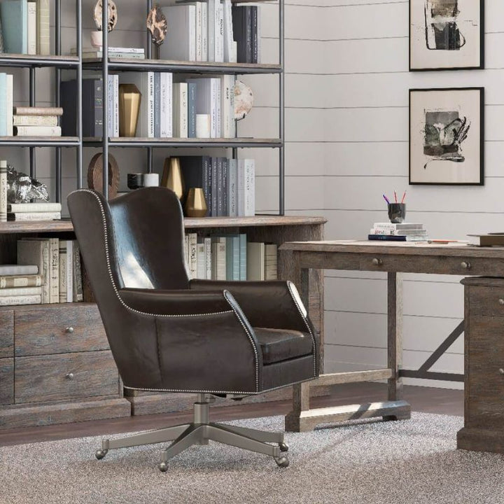 A stylish home office exudes sophistication with a dark brown aniline leather Plaza 66 Office Swivel Chair by Fairfield Chair, featuring sleek metal legs. The setup includes a wooden desk with drawers, a large bookcase filled with books and decorative items, framed artwork on the walls, and a neutral-toned carpet covering the floor.