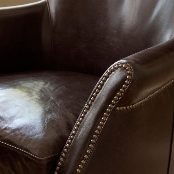 A close-up of the Plaza 66 Office Swivel Chair by Fairfield Chair showcases the texture of its dark brown aniline leather and silver-colored decorative nailhead trim along the edge of the armrest. The seating surface appears slightly worn, reflecting light softly, and adds a sophisticated touch to this elegant piece.