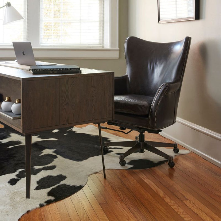 A stylish home office with a wooden desk featuring decorative items and an open laptop. The sophisticated touch of the Plaza 66 Office Swivel Chair from Fairfield Chair, crafted in black aniline leather, complements the hardwood flooring and black-and-white cowhide rug. Sunlight filters in through the window.