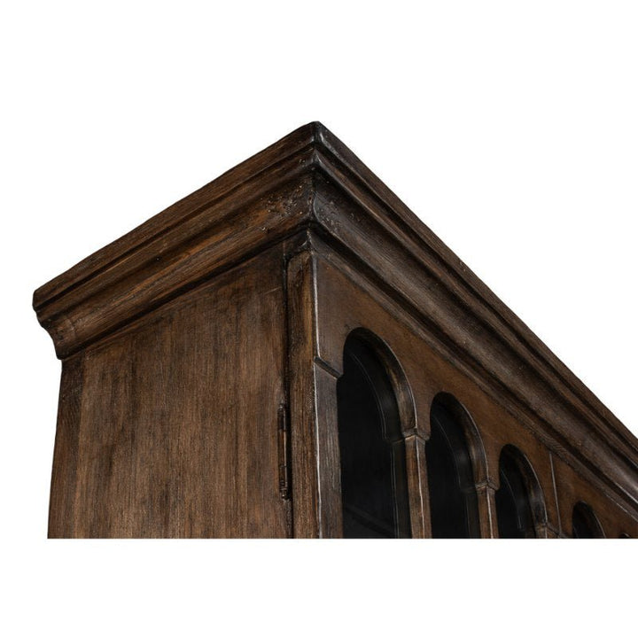 Close-up of the upper corner of a dark wooden Refined Arches Tall Bookcase by Sarreid, featuring a cornice and a series of arched glass-paneled doors. The wood has a slightly weathered appearance, showcasing its rustic, vintage design.