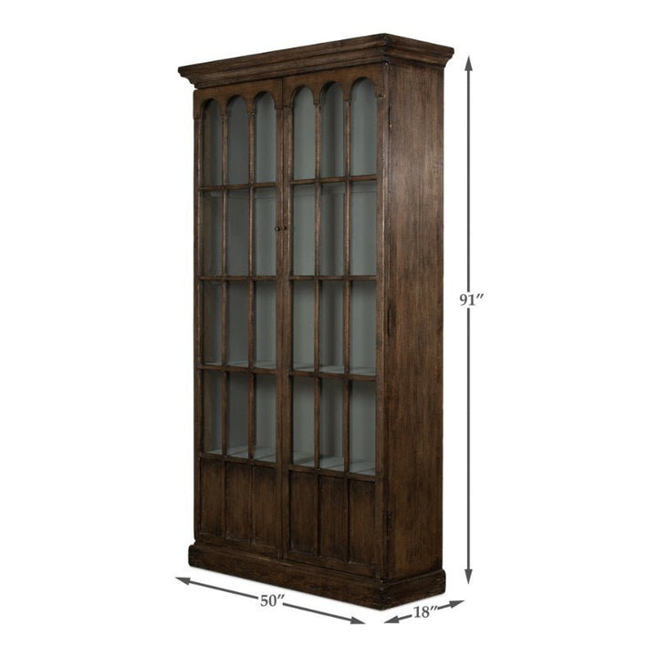 The Refined Arches Tall Bookcase by Sarreid is a tall, dark wood cabinet with glass-paneled double doors and elegant arches. Standing at 91 inches high, 50 inches wide, and 18 inches deep, this traditional style bookcase features a classic antique design perfect for displaying items or providing ample storage.