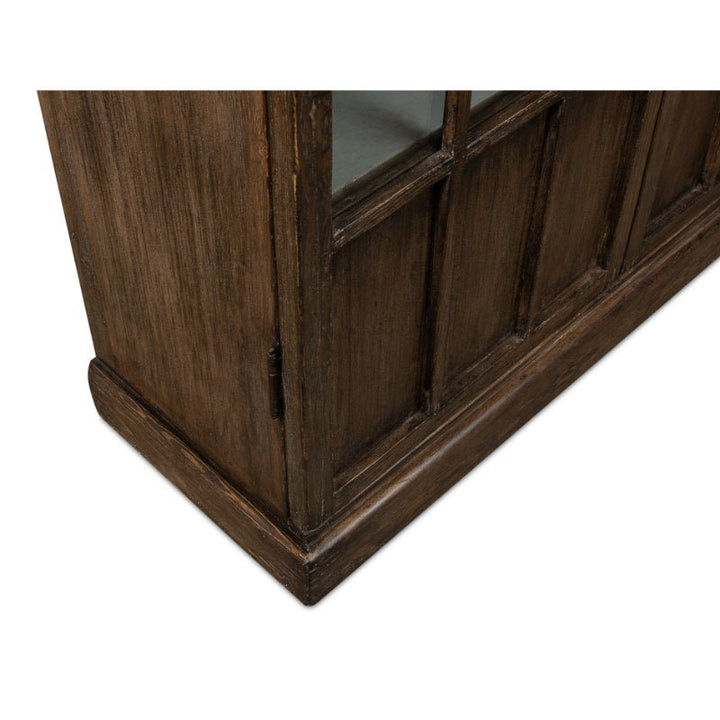 A close-up view of the base and side of the Refined Arches Tall Bookcase by Sarreid reveals its traditional style with a slightly weathered finish. The cabinet features glass-paneled doors with an antique design and hinges. The wood has a dark, rustic appearance highlighted by the texture and grain, offering ample storage for your needs.