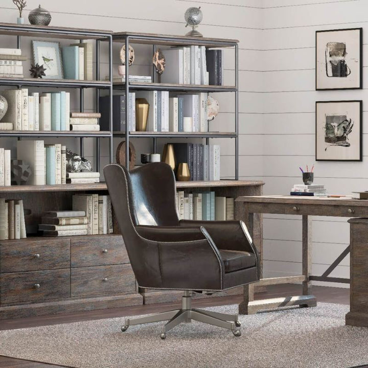 A home office with rustic elegance, showcasing wooden furniture and a large bookshelf stocked with books and decorative items. In the foreground, there's a dark brown leather swivel chair positioned in front of a wooden desk next to the Rustique Lateral File Cabinet by Fairfield Chair. The floor is covered with a light-colored rug.