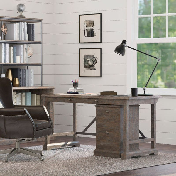 A cozy home office showcases a Rustique Writing Desk and Mobile File from Fairfield Chair, complemented by a black adjustable desk lamp, a cushioned office chair, and shelves brimming with books and decor items. The room is bathed in natural light from a large window, and two framed artworks elegantly adorn the wall above the desk, highlighting rustic charm.