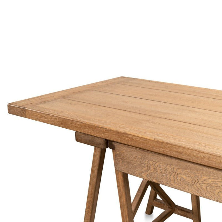 A Sarreid Sawhorse Desk in Natural Polished Old Pine, radiating rustic charm with its rectangular design and trestle legs. The desk boasts a light brown finish that beautifully highlights the natural wood grain of reclaimed pine. Only part of the tabletop and legs are visible in the image.