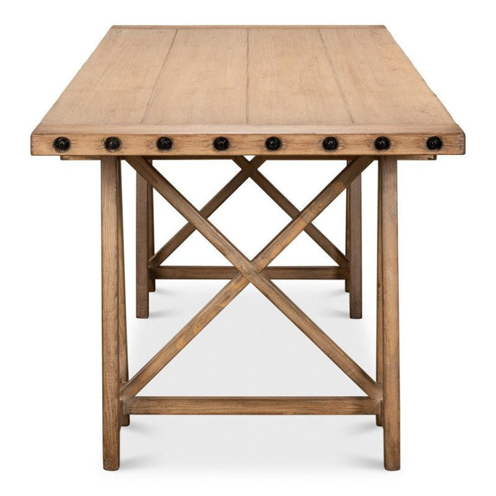 A rectangular wooden table named "Sawhorse Desk, Natural Polished Old Pine 87"" by Sarreid is shown from the front. It features exposed drawer pulls along the edge and an X-shaped support structure connecting the legs at the base. The desk exudes rustic charm, reminiscent of a traditional sawhorse desk in reclaimed pine.