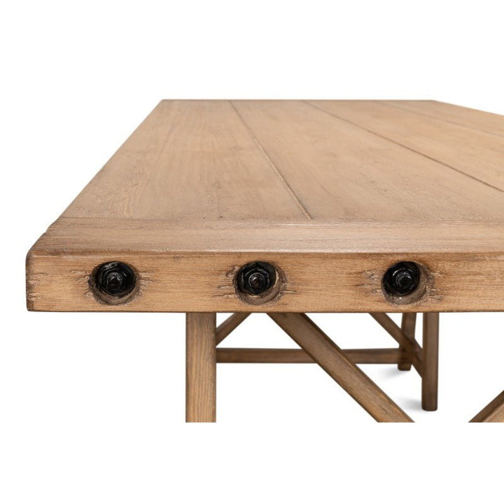 A close-up view of the Sarreid Sawhorse Desk, crafted from natural polished old pine, showcases its rustic charm and industrial design. Measuring 87 inches, this table features three round black bolt details on the edge and crisscrossed legs that provide sturdy support, reminiscent of a classic sawhorse desk.