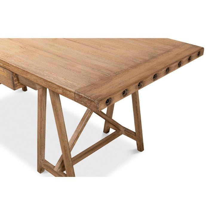 The Sarreid Sawhorse Desk features a rustic design with a natural polished old pine rectangular top, showcasing visible wood grain. The desk’s X-shaped leg supports are reminiscent of traditional sawhorse desks, complemented by circular metal details along the edges. Its light brown finish enhances its rustic charm, completing the look of this 87" piece.