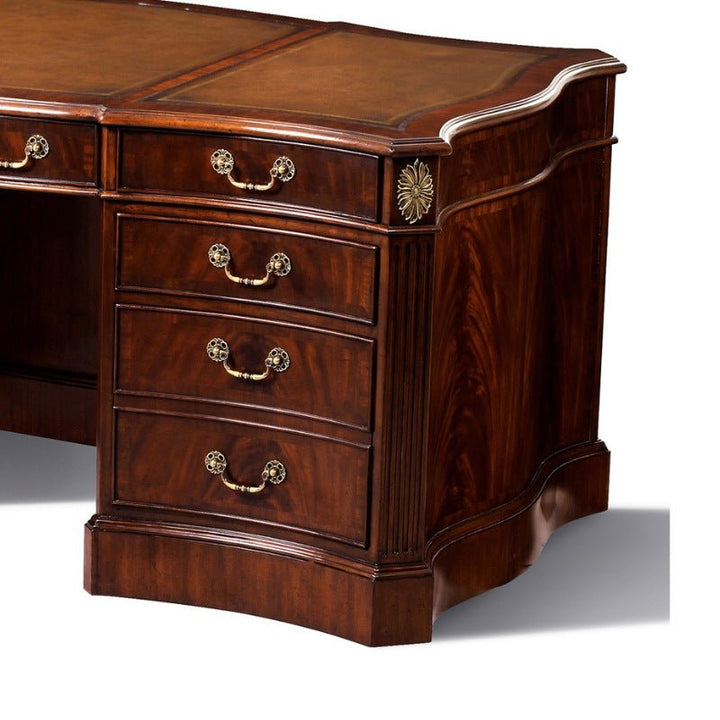 The Serpentine Writing Desk by Aston Court features a polished, dark wood finish and intricate metal handles adorned with decorative floral motifs on the drawer pulls. This elegant desk includes four spacious drawers on one side, classic fluted columns, and a wavy-edged design on the opposite side, all complemented by an antique brown leather top.