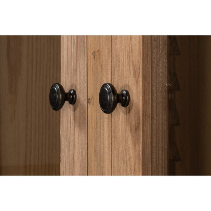 Close-up of two black round knobs on a light wood cabinet door with a natural brushed finish. The wood has a vertical grain pattern, and the knobs have a simple, classic design. The right side of the image shows the corner of the cabinet door and part of the grooved interior shelf from the Tanner Bookcase in Natural by Sarreid.