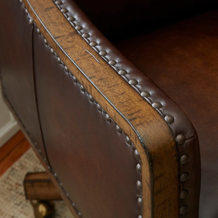 Close-up of The Shard Office Swivel Chair by Fairfield Chair, featuring Aniline leather upholstery with a wooden frame and metal studs along the edges. The chair is positioned on a light-colored woven rug.