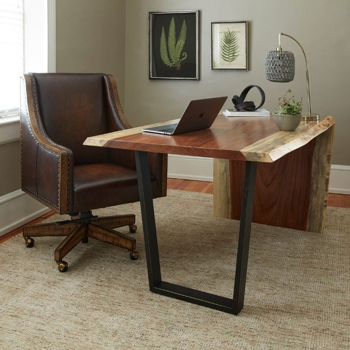 A home office features a polished wooden desk with a live edge, black metal legs, a laptop, a small plant, and decorative items. Adjacent to the desk is The Shard Office Swivel Chair from Fairfield Chair on casters. Fern artwork hangs on the wall above the desk, adding a natural element.