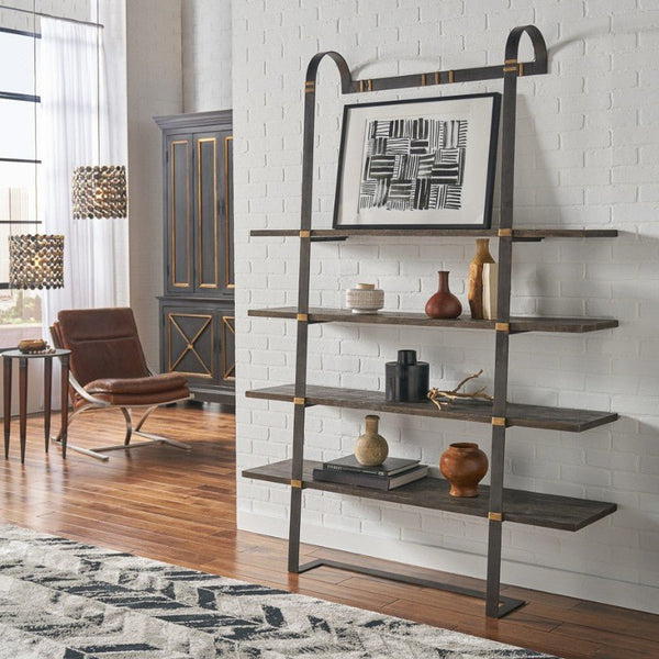 A modern living space features The Shepard's Crook Bookshelf by Sarreid, showcasing dark acacia wood shelves supported by a metal frame, perfect for displaying decorative items. Atop the shelf sits a minimalist black-and-white artwork. Nearby, a brown leather chair, a side table, and a patterned rug contribute to the contemporary elegance.