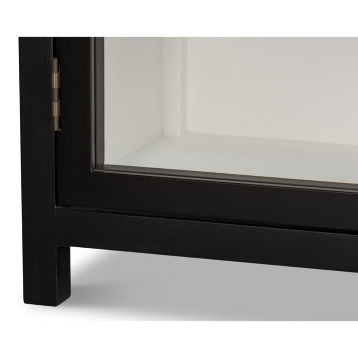 Close-up of the Walker Bookcase, Black | Sarreid, showcasing a black-framed glass display cabinet corner with a brushed black exterior. The hinge is visible on the left side, and the neat, minimalist design accentuates the smooth, sleek edges. The interior is white, enhancing the transparency and clarity of the glass panel.