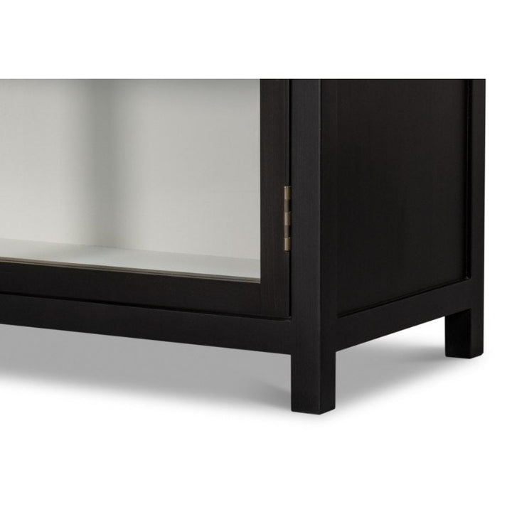 The image displays the lower portion of the Walker Bookcase in Black by Sarreid, featuring glass doors. This cabinet, showcasing a rustic elegance bookcase design, boasts clean lines and short legs. Its brushed black exterior offers a striking contrast to the light-colored interior, resulting in a simple yet sophisticated appearance.