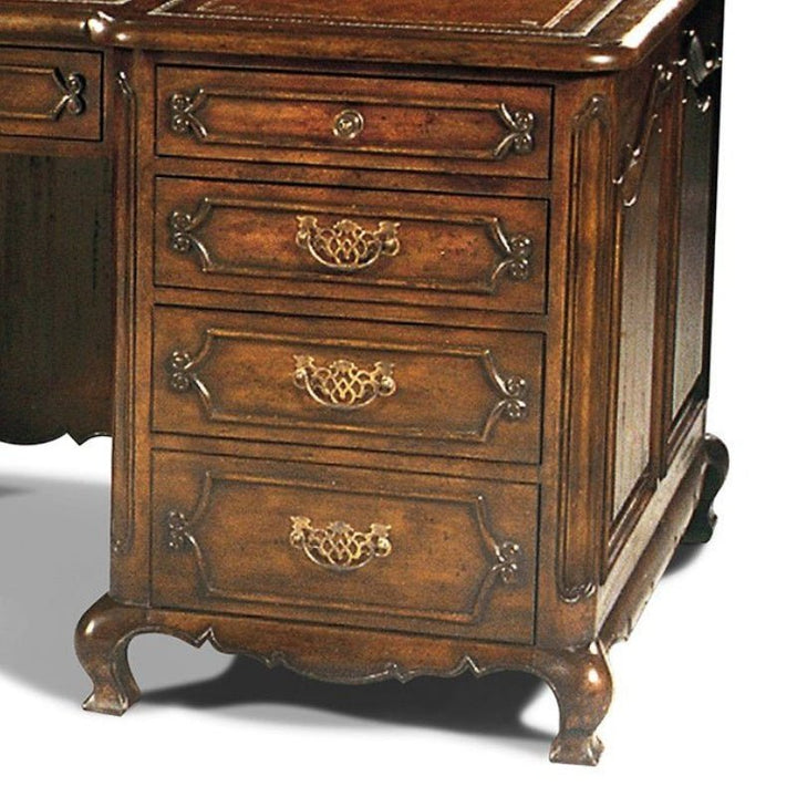 The Wesley Writing Desk by Aston Court is a vintage wooden piece with intricate carvings and ornate brass handles. This handcrafted desk features four drawers, each adorned with decorative pulls, and stands on curved legs. The polished wood boasts a rich brown finish complemented by an antique brown leather top.